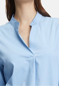 Jasnoniebieska bluzka z kołnierzykiem i rozciętym dekoltem, charakteryzująca się subtelnym detalem w postaci pliski oraz gładkim, teksturowanym materiałem.