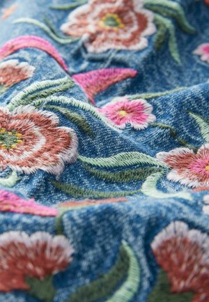 Tissu en denim bleu avec des fleurs roses et orange brodées et des feuilles vertes, montrant une texture et des coutures détaillées.