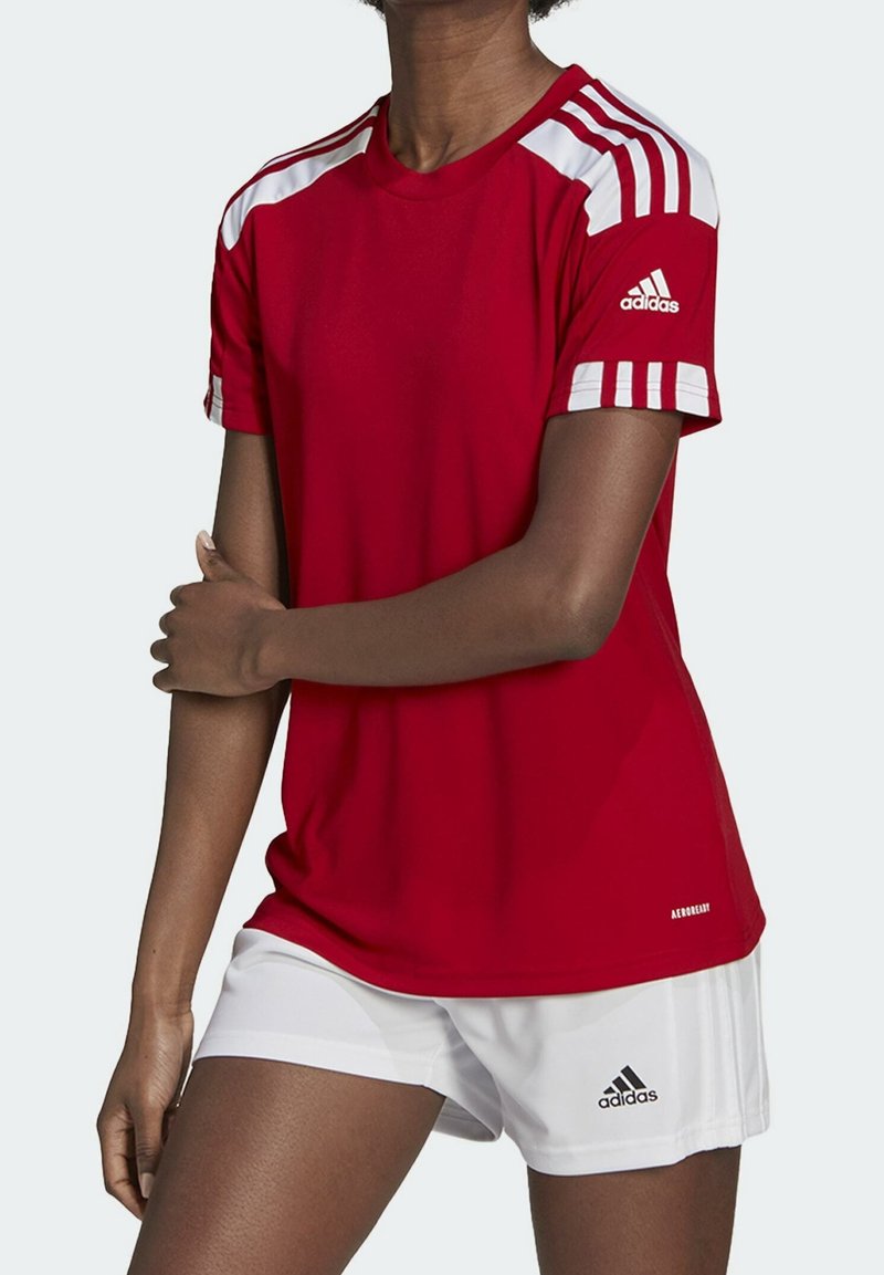 Personne portant un maillot de sport rouge Adidas avec des bandes blanches et un short Adidas blanc, debout avec un bras plié sur le corps.