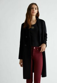 Cappotto di lana nero con due bottoni, maniche lunghe e taglio dritto, abbinato a pantaloni bordeaux e a una maglietta nera con un dettaglio del logo.