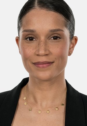 Gold necklace with small circular pendants on a delicate chain, worn with a black blazer and a neutral background. Smooth metal finish.