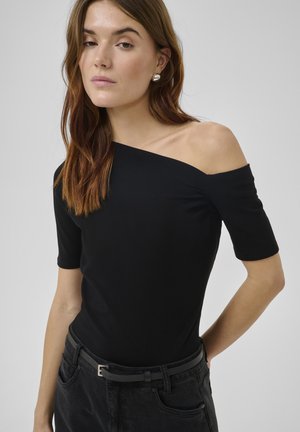 Donna con capelli castani lunghi indossa una maglietta nera con spalle scoperte, cintura nera e pantaloni neri davanti a uno sfondo grigio semplice.