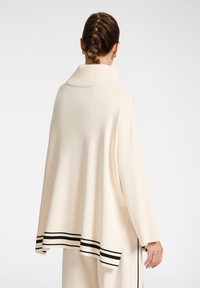 Poncho de color crema con una silueta amplia y caída, que cuenta con un collar doblado y acentos de rayas negras a lo largo del dobladillo. Tejido suave y texturizado.