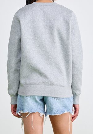 Personne portant un sweat-shirt gris uni et un short en jean bleu clair effiloché, vue de dos sur un fond uni.