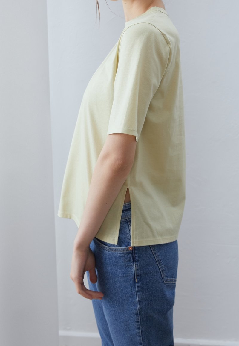 Personne portant une chemise jaune clair à manches courtes avec une fente latérale et un jean bleu, vue de profil sur un fond de mur uni.