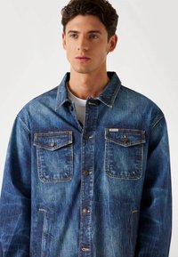 Denim jacket in faded blue with a button-down front, two chest pockets, and a branded label. Relaxed fit with a classic collar and textured fabric.