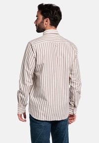 Man met donker haar en een baard, gekleed in een beige en witte verticaal gestreepte lange mouwen shirt en blauwe spijkerbroek, met zijn rug naar de camera, tegen een witte achtergrond.