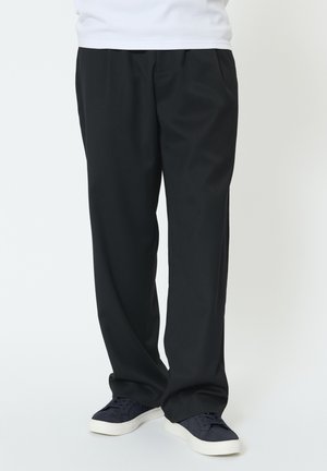 Person iført løstsiddende sorte bukser og marineblå sneakers med hvide såler, stående mod en ensfarvet lys baggrund.