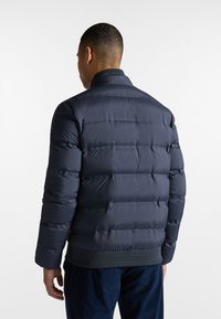 Marineblauwe gewatteerde jas met horizontale baffels, elastische manchetten en een geribde zoom. Voorzien van een hoge kraag en een gladde textuur.