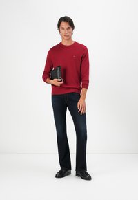 Pull en tricot rouge, jean en denim foncé et chaussures noires. Modèle tenant une pochette noire, avec un fond blanc uni.