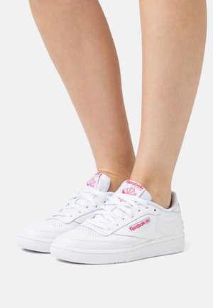 Bílé kožené tenisky s růžovým logem a akcenty, kulatá špička, perforovaný design pro ventilaci a ploché gumové podrážky.