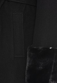 Manteau en laine noire avec un poignet en fausse fourrure douce. Dispose d'une poche latérale et d'un design épuré et ajusté. La texture semble douce et lisse.