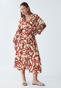 Duga haljina s uzorkom u smeđoj i krem bojama, s remenom za vezanje, kopčanjem sprijeda na gumbe i širokim rukavima. Nošena s crno-bijelim ravnim sandalama.