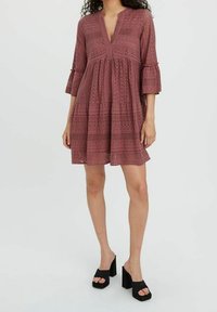 Robe rose froncée avec un décolleté en V, des manches trois-quarts et des motifs en dentelle. Portée avec des talons compensés noirs.