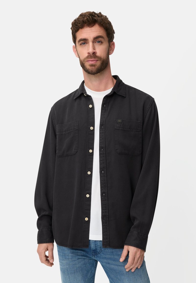 Chemise noire à manches longues en coton, avec deux poches poitrine et des boutons de couleur claire. Portée par-dessus un T-shirt blanc, avec une coupe décontractée.