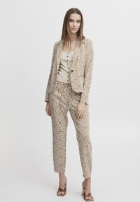 Braunes und weißes gemustertes Anzugensemble mit tailliertem Blazer und passenden Hosen, dazu ein strukturiertes beiges Oberteil und braune Absatzsandalen.