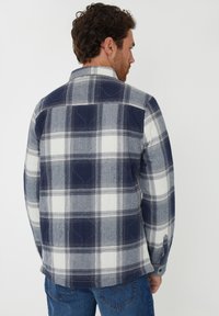 Koszula flanelowa w kratę w kolorze granatowym, białym i szarym. Charakteryzuje się wzorem w kratę, zapięciem na guziki i zaokrąglonym dołem. Luźny krój z długimi rękawami.