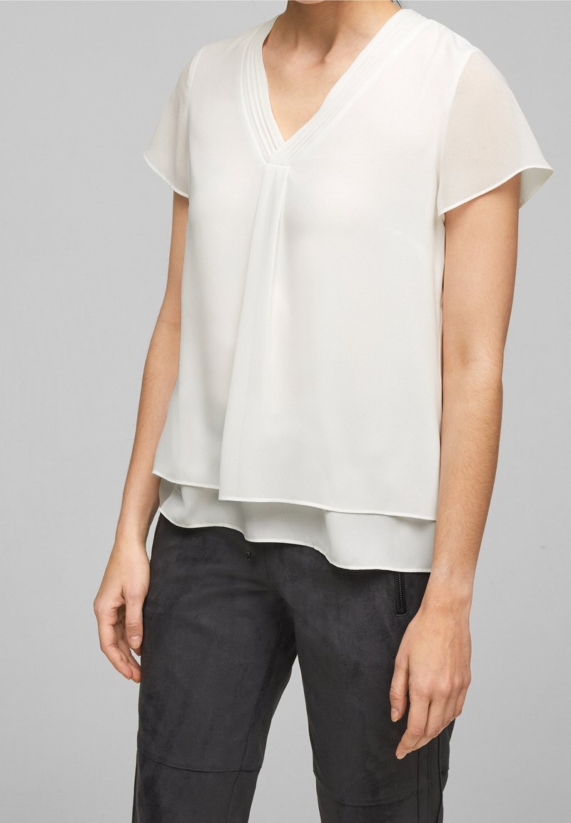 Femme portant un chemisier blanc à manches courtes superposé et un pantalon texturé foncé, debout devant un fond gris uni.