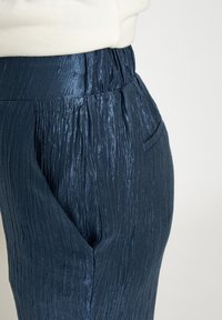 Donkerblauwe broek met gerimpelde textuur, elastische tailleband en zijzakken. De stof heeft een subtiele glans en een gestructureerd ontwerp.