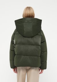 Person med kort blondt hår iført en oversize mørkegrønne puffjakke med hætte, stående foran en ensfarvet lys baggrund.