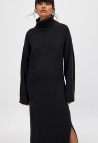 Abito nero lavorato a maglia con alto collo a tromba, maniche lunghe e spacchi laterali, caratterizzato da un tessuto testurizzato e una vestibilità comoda.