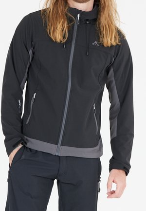 Homme portant une veste softshell noire et grise zippée avec des poches zippées et de longs cheveux blonds, debout avec une main dans la poche.