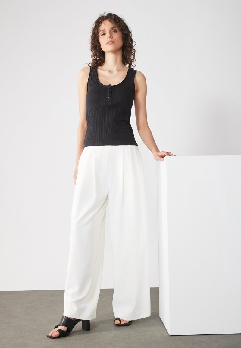 Black ribbed tank top with button detail, paired with white wide-leg trousers and black heeled sandals. Simple, well-defined shapes and textures.