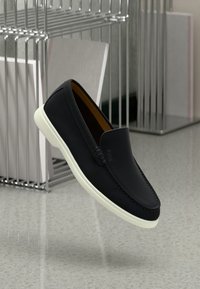 Sorte slip-on sko med hvit såle og "BOSS"-logo, plassert midt i luften over en flekkete grå gulv og en metalltrådorganisator i bakgrunnen.