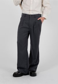 Homme portant un pantalon large gris foncé, une chemise boutonnée rayée crème rentrée dans le pantalon, une main dans la poche et des chaussures noires.