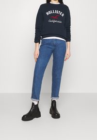 Marinblå sweatshirt med "Hollister California"-tryck, ihop med ljusblå denimjeans med uppvikta ärmar och svarta ankelstövlar.