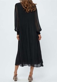 Svart, flödande maxiklänning med långa genomskinliga ärmar, plisserat tyg och smockade ärmslut. Har en avslappnad silhuett och är knälång.