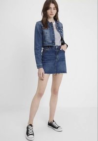 Chaqueta de mezclilla corta con dos bolsillos en el pecho, combinada con una falda de mezclilla de cintura alta, que presenta un dobladillo deshilachado. Zapatillas de lona negras completan el look.