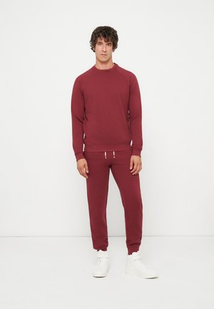 Homme debout contre un mur blanc, portant un sweat-shirt bordeaux assorti et un jogging avec des baskets blanches, faisant face avec une expression neutre.