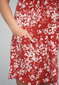 Punainen mekko, jossa on valkoisia ja vaaleanpunaisia kukkia sisältävä kukkakuosi, vyötärö kapeana ja sivutaskut. Teksturoidusta kankaasta, jossa napin yksityiskohdat.