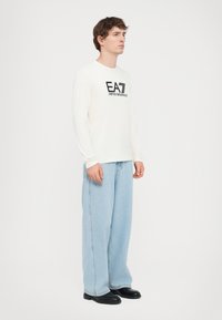 Langermet hvit T-skjorte med svart trykket logo, kombinert med lyseblå vide jeans og svarte sko. Enkelt, uformelt antrekk.