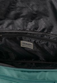 Forro interior negro de una bolsa con un patrón hexagonal, con una etiqueta cosida que dice "JASLEN EST. 1987" y un bolsillo con cremallera color verde azulado.