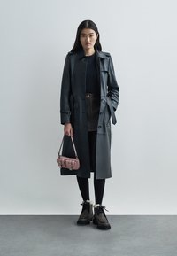 Schwarzer Leder-Trenchcoat mit einem taillierten Gürtel, kombiniert mit einem dunklen Rock, schwarzen Leggings und robusten Stiefeletten. Hält eine pinke Handtasche.