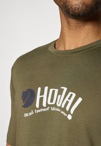 Olīvzaļš kokvilnas T-krekls ar grafisko apdruku, kurā redzams tumši zilas krāsas zirga siluets un balts teksts "HOJA! CYCLING EQUIPMENT FJÄLLRÄVEN SVERIGE."