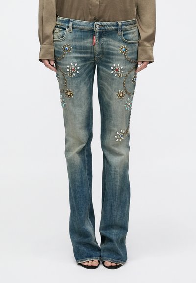 Blaue ausgewaschene Jeans mit ausgestellten Beinen, verziert mit mehrfarbigen Perlen in Blumenmustern, getragen mit einem beigen Langarmshirt und schwarzen offenen Schuhen.