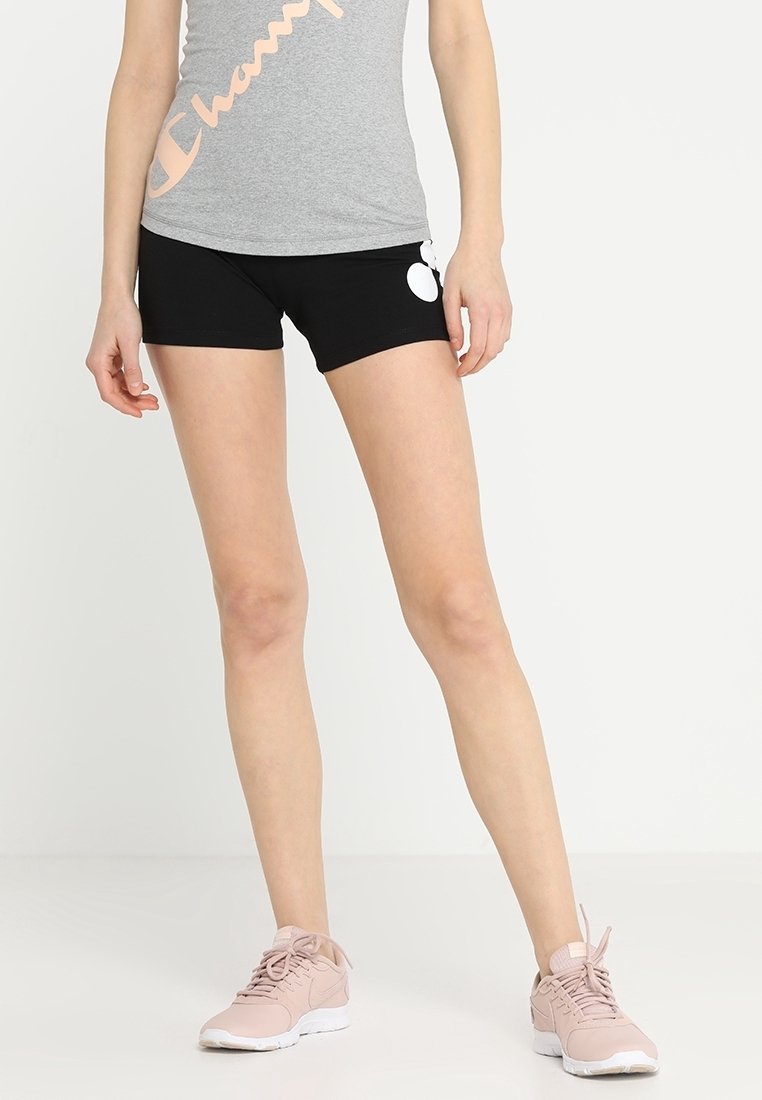 Femme portant un short de sport noir avec un logo blanc, un débardeur gris Champion et des baskets rose clair, debout devant un fond blanc.