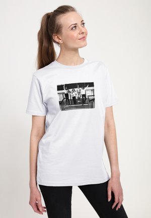 Kvinde med hestehale iført hvid t-shirt med sort-hvidt foto af fire personer, der hopper på udendørs trapper.