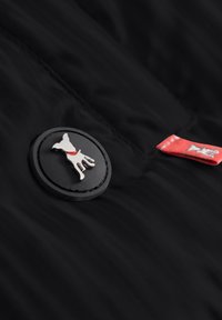 Schwarzer Stoff mit einem runden Aufnäher, der die Silhouette eines Hundes in Silber und ein rotes Halsband zeigt. Rotes Akzentetikett auf dem Material in der Nähe des Aufnähers.