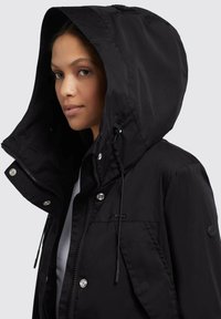 Veste à capuche noire en tissu léger. Elle présente une fermeture éclair, des boutons-pression et une capuche avec cordon de serrage. Accentuée par un détail de logo.
