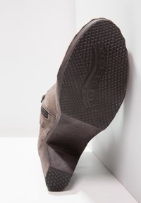 Brown suede ankle boot with a textured rubber sole, featuring a geometric heel and a zip detail on the side. Sole has a ridged pattern.