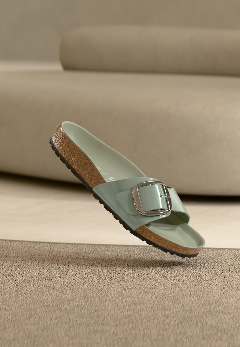 Πράσινο σανδάλι τύπου slip-on με πάτο από φελλό και μεγάλο ασημένιο αγκράφα, τοποθετημένο πάνω σε υφασμάτινη μπεζ επιφάνεια με θολό μπεζ φόντο.