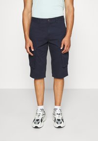 Shorts cargo marine avec deux grandes poches latérales, fermeture à bouton et coupe décontractée, associés à des baskets blanches et noires.