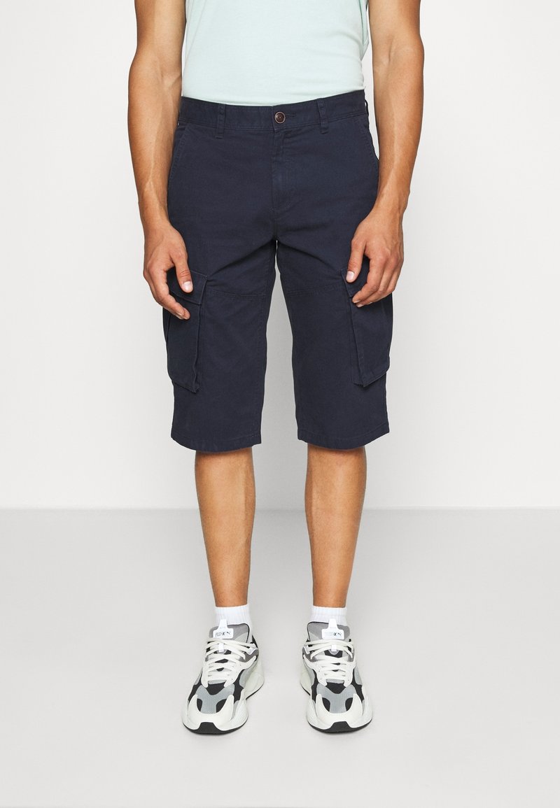 Shorts cargo marine avec deux grandes poches latérales, fermeture à bouton et coupe décontractée, associés à des baskets blanches et noires.