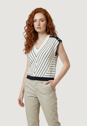 Jeune femme aux longs cheveux roux bouclés portant un pull sans manches blanc à rayures noires et un pantalon beige, debout avec une main dans la poche.