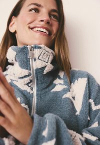 Sininen fleece-takkia, jossa on valkoisia pilvikuosisia, korkea kaulus ja hopeisia vetoketju yksityiskohtia. Pehmeä tekstuuri näkyy kankaassa.