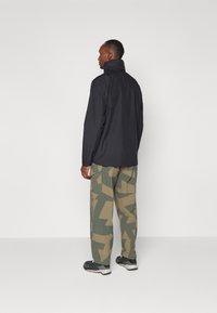 Black jacket with a high collar, paired with camouflage-patterned pants in green and beige, worn with dark athletic shoes.